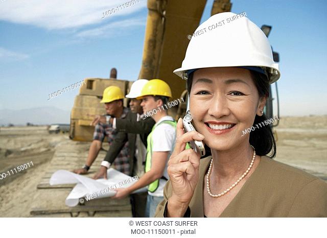 Surveyor in hard hat standing in front of heavy machinery Using Cell Phone on Construction Site