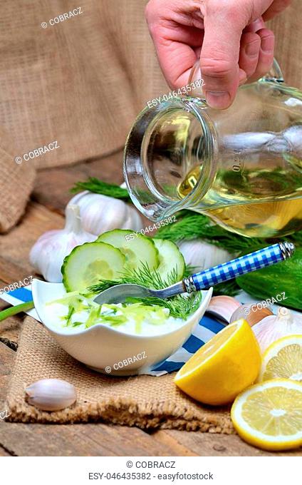 Man pours oil into Tzatziki - traditional Greek dressing or dip sauce, garlic, lemon, dill, cucumber, jug with oil, blue spoon and decoration in background