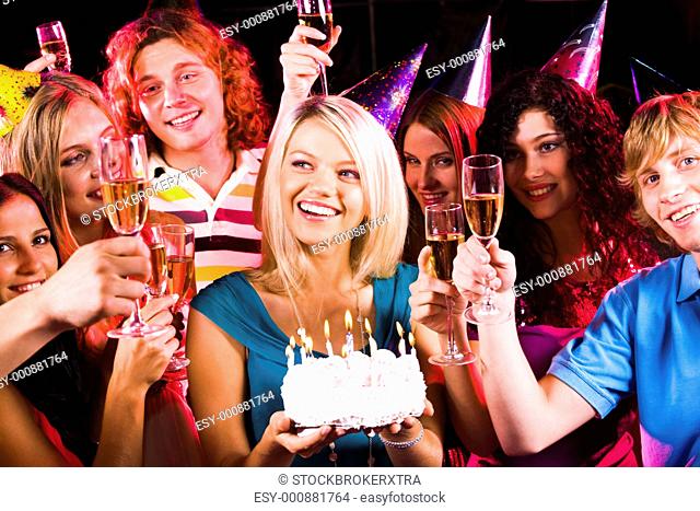 Portrait of joyful girl holding birthday cake surrounded by friends with flutes of champagne