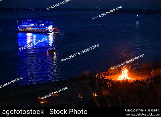16 April 2022, Hamburg: The blue illuminated paddle steamer Louisiana Star sails along the Elbe behind one of the smaller Easter bonfires in Blankenese