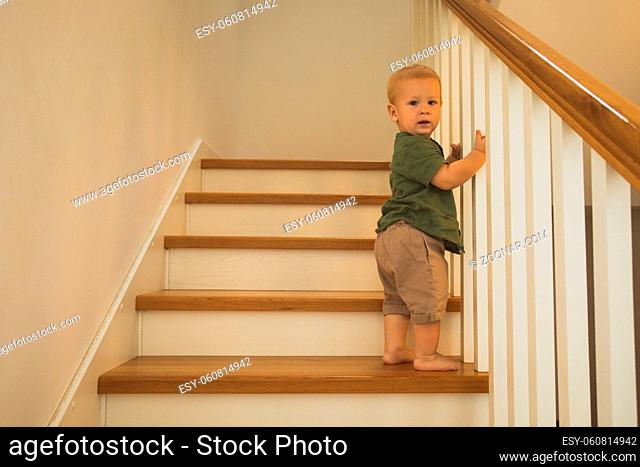 Cute toddler boy, barefoot in green t-shirt standing on stairs at home, holding wooden railings, looking at the camera. New achievents and skills of little boy