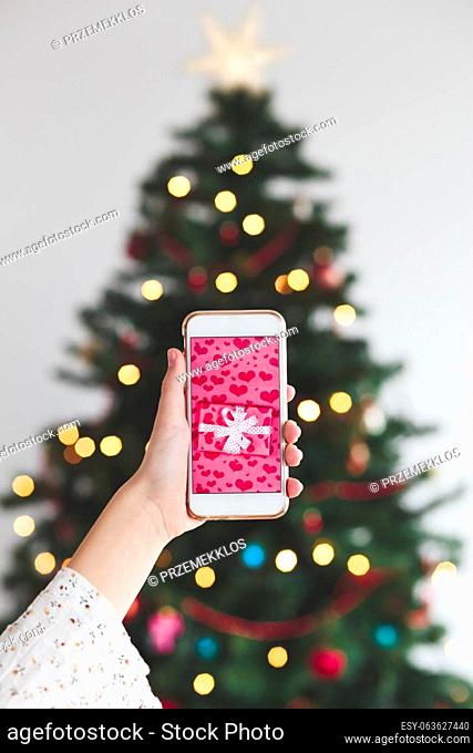 Girl holding the smartphone with wrapped gift on the screen and christmas tree in the background