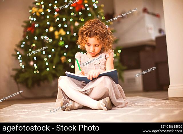 Christmas mood photo of cute curly girl, sitting next to festive tree and drawing in her sketch book. Family winter holidays concept