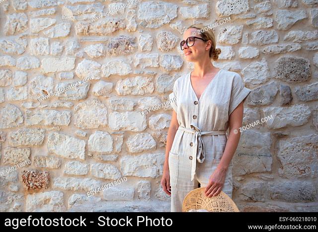 Portrait of beautiful cheerful blonde woman wearing one piece sundress and summer hat, standing in front of old medieval stone wall