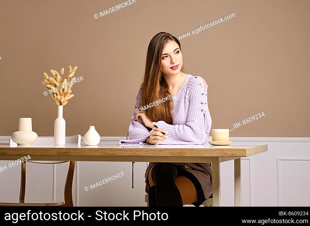 Pensive young woman sits behind the table and thinking about what to write in postcard