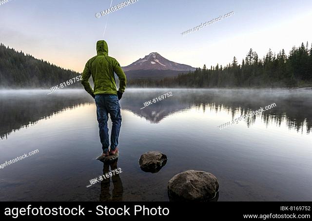 Young man standing on stone, reflection of Mt. Hood volcano in Trillium Lake, at sunrise, Oregon, USA, North America