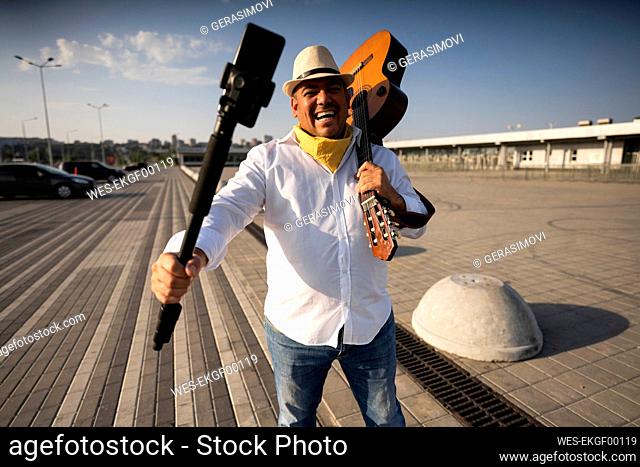 Happy street musician holding guitar taking selfie through mobile phone on tripod