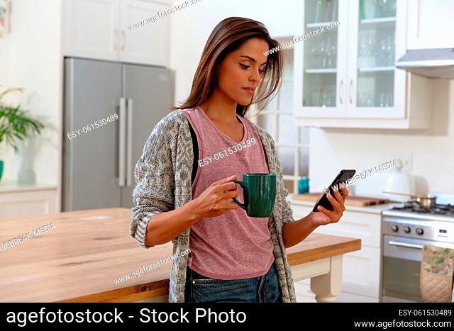 Caucasian woman standing in kitchen drinking cup of coffee using smartphone and smiling