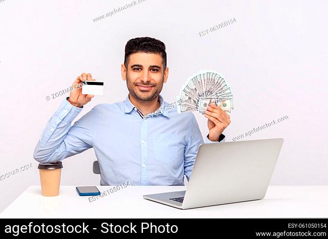 Bank loan, electronic money. Positive businessman sitting office workplace with laptop, holding dollars and credit card, looking at camera with smile