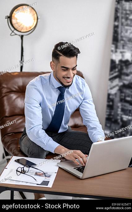 Good mood. Smiling elegant young male working on laptop sitting in leather chair in room during daytime