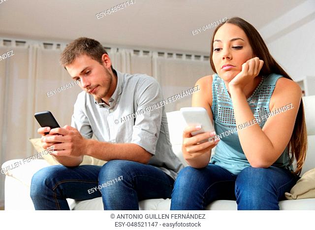 Bored couple online with their smart phones sitting on a couch in the living room at home