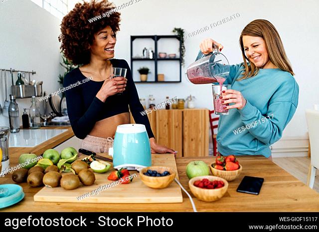 Happy young woman looking at friend pouring smoothie in drinking glass standing at table