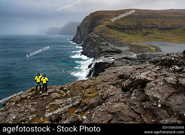 Couple of man in yellow jackets stands on the rocky cliff on the seaside on the foggy horizon background on Faroe Islands. Shoot from the back