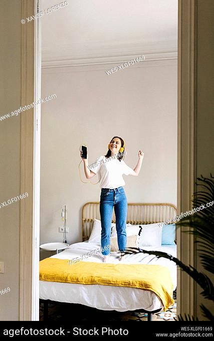 Cheerful young woman with mobile phone wearing headphones while dancing on bed at home
