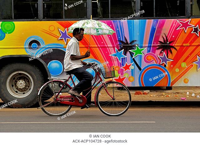 man on a bicycle with an umbrella with colourful bus, Chidambaram, Tamil Nadu, India