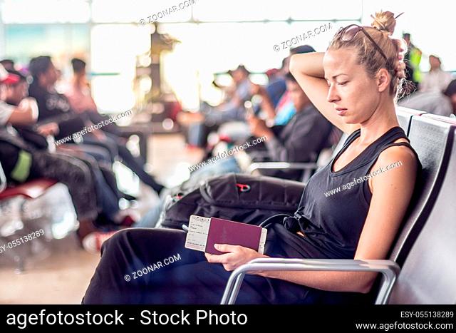 Casual tanned blond female traveler holding cell phone, passport and boarding pass while waiting to board a plane at the departure gates at the asian airport...