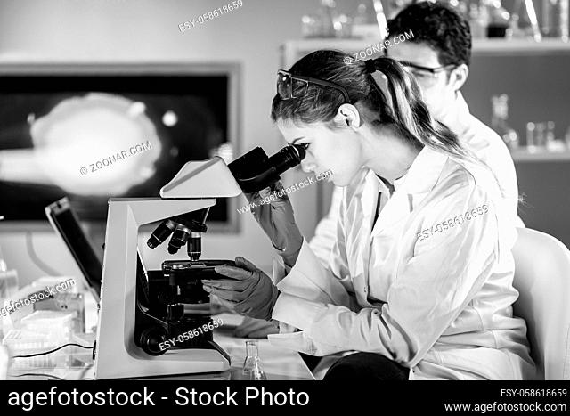 Life scientists researching in laboratory. Attractive female young scientist and her post doctoral supervisor microscoping in their working environment