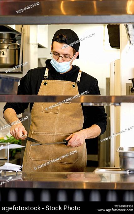 Chef with protective face mask sharpening knifes in kitchen