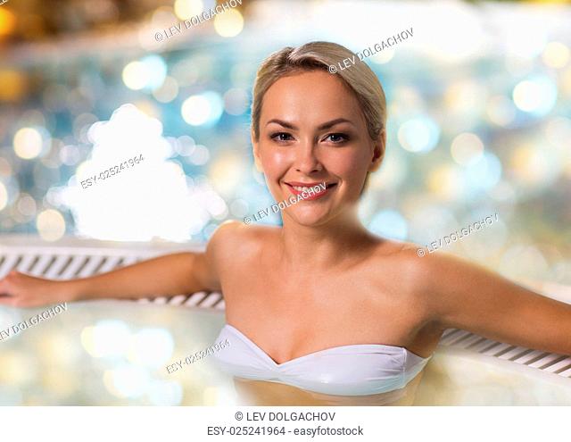 people, beauty, spa and relaxation concept - beautiful young woman wearing bikini swimsuit sitting in jacuzzi at poolside over holidays lights background