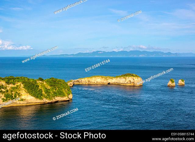 Indonesia. Strait between the islands. Day. Rocky coast with jungle and two small islets. Mountains and clouds on the horizon