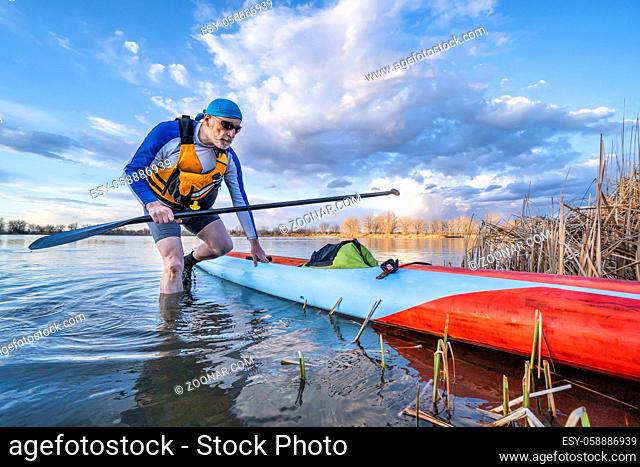 senior male paddler with his stand up paddleboard on a shore of calm lake, solo paddling as fitness and training with social distancing