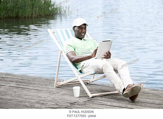 Man relaxing on deck chair using digital tablet by lake