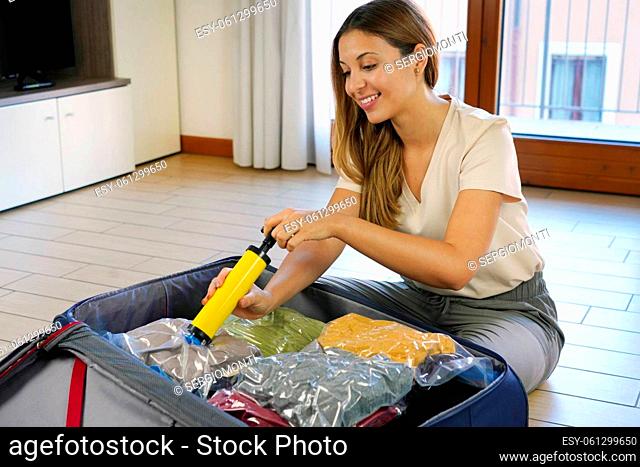 Attractive smiling woman using vacuum pump saving space in her luggage sitting on floor. Vacuum clothing storage compressed package