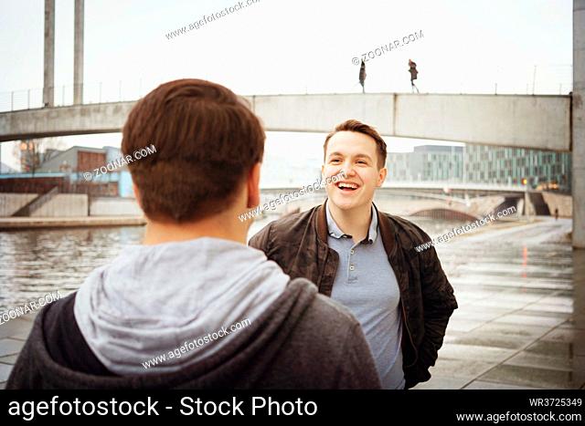 two male teenage friends having a fun conversation - lifestyle or city life concept - urban riverside location in Berlin Germany