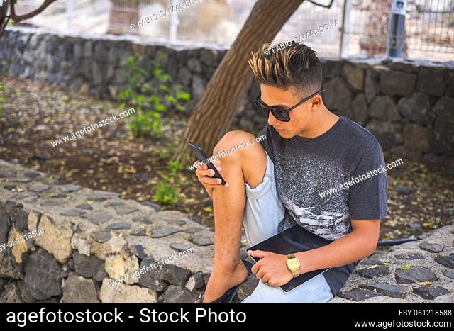 Young caucasian boy use mobile phone cellular outside at the garden sitting and relaxing - online new generation people connected to internet and social life