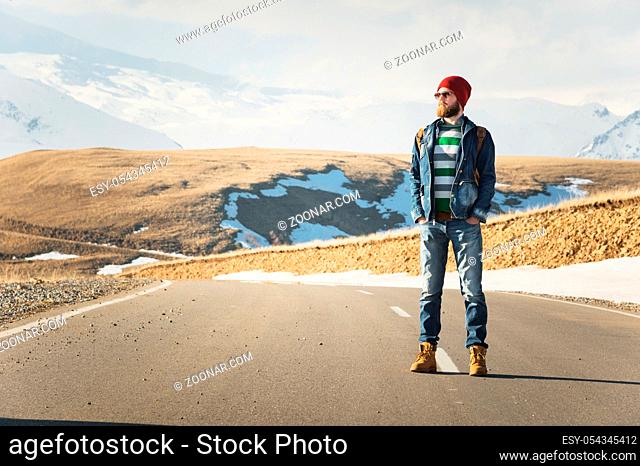 A stylish bearded hipster in sunglasses with a vintage backpack stands on a country road asphalt on a sunny day. The concept of hitchhiking and hiking