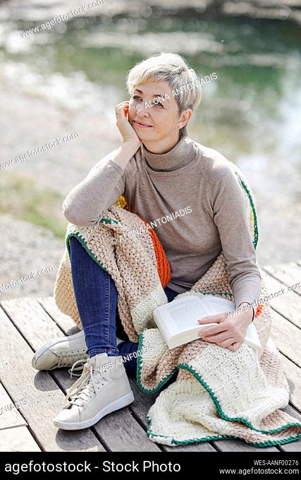 Thoughtful woman sitting with book and knit blanket at pier