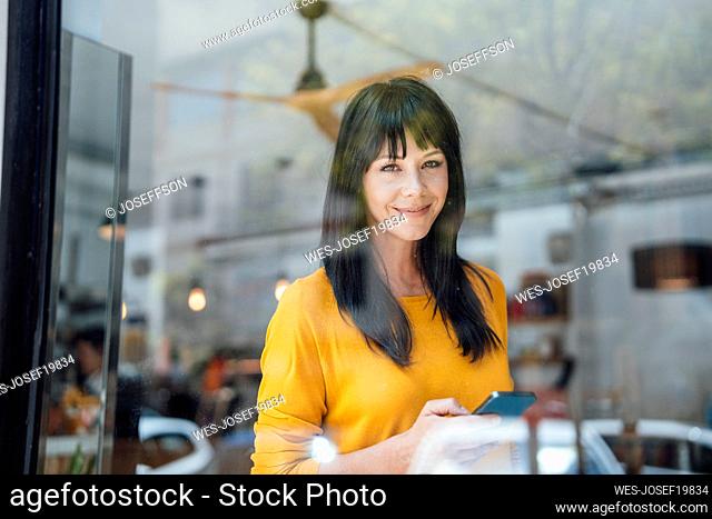 Smiling mature woman with smart phone seen through glass in cafe