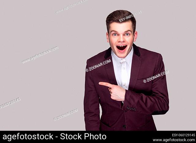 Portrait of surprised handsome young man in violet suit and white shirt, standing, looking at camera and pointing at copy space with amazed face