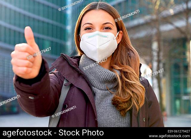 Close up of optimistic business woman wearing protective mask KN95 FFP2 showing thumbs up in modern city street and looking at camera