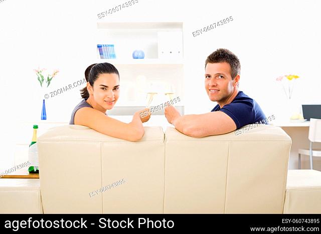 Love couple clinking champagne glasses at home on sofa. Smiling and looking at camera