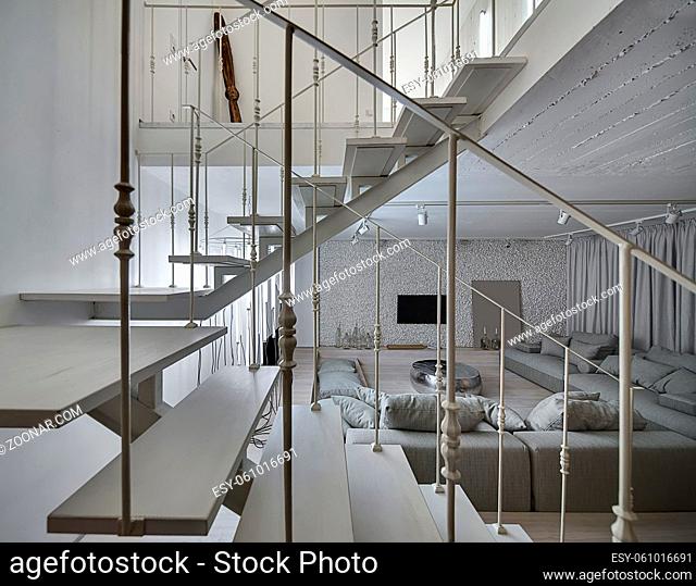 Interior in a modern style with light walls and a parquet on the floor. There is a white wooden stair with metal railing, gray sofas, sparkle pouf