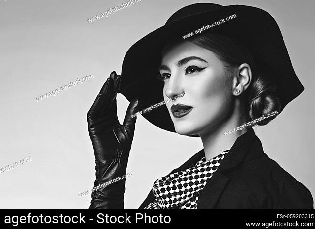 Beautiful young woman with red lips make-up in retro black and white dress, hat, jacket and leather gloves standing on grey background. Monochrome