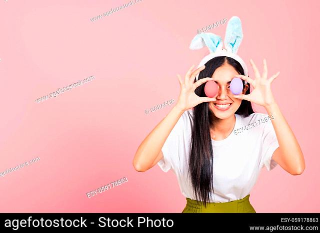 Happy beautiful young woman smiling wearing rabbit ears holding colorful Easter eggs front eyes, Thai female with bunny ear hold easter egg covering eye