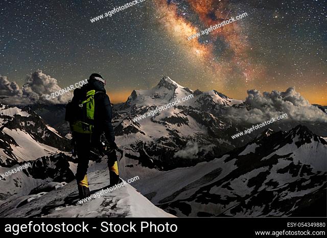Night photo. Professional mountaineering guide fully equipped and with a backpack on his shoulders and an ice ax in his hands stands on top of the mountain in...