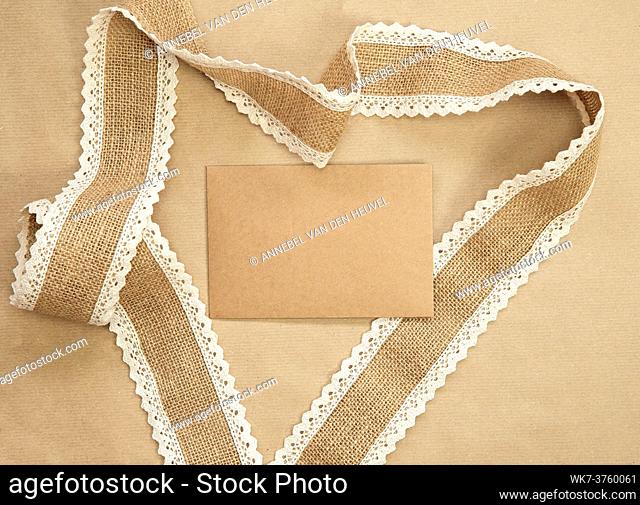Flat Lay composition with blank brown plain greeting card surrounded with natural burlap ribbon on craft paper background texture, top view design