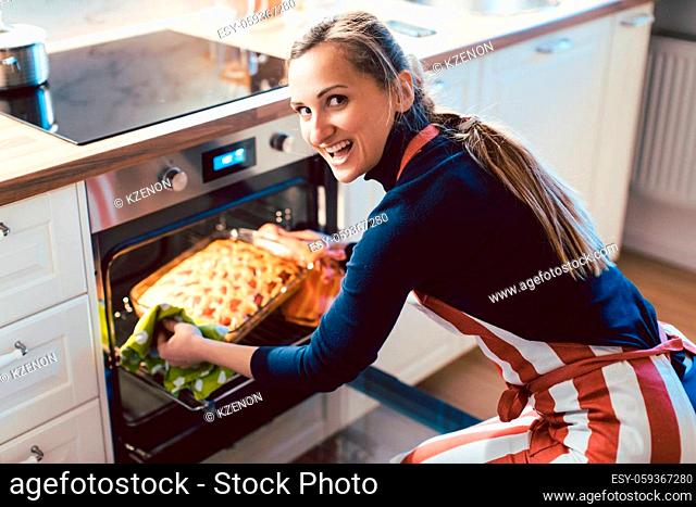 Portrait of cheerful young female wearing apron removing freshly baked apple pie from oven in kitchen at home