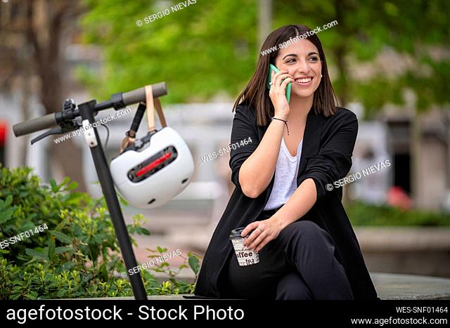 Young smiling businesswoman talking on mobile phone while sitting by electric push scooter