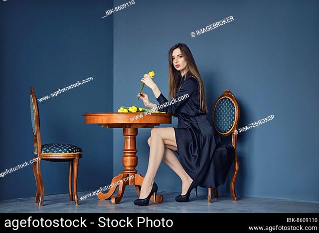 Beautiful young woman sits behind the table in victorian style with yellow tulips in hand