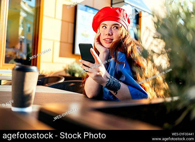 Young woman in beret hat day dreaming while holding mobile phone at sidewalk cafe