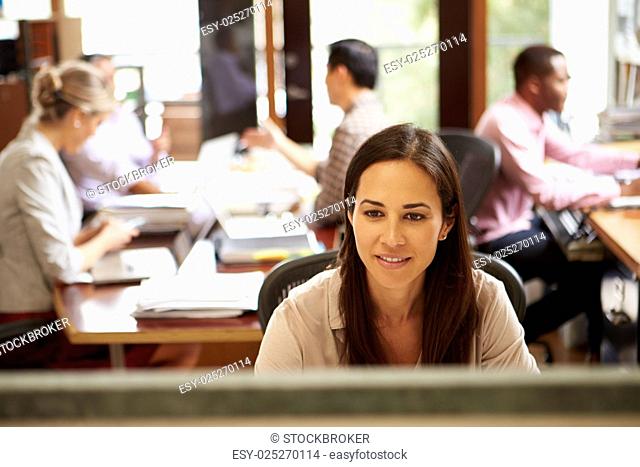Businesswoman Working At Desk With Meeting In Background