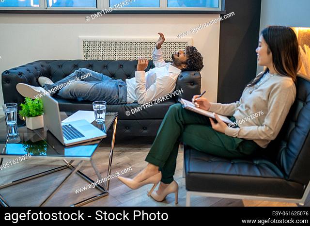 Focused qualified psychologist listening to complaints of a young man lying on the couch in her office