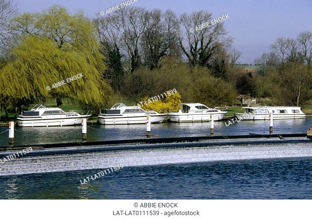 River Thames. Water. Row of four white boats moored by river bank. Evenly spaced out. Narrow boat. Trees. Willows. ranches being blown by wind