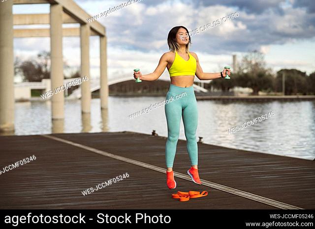 Cheerful sportswoman jumping with dumbbells on jetty at lakeside