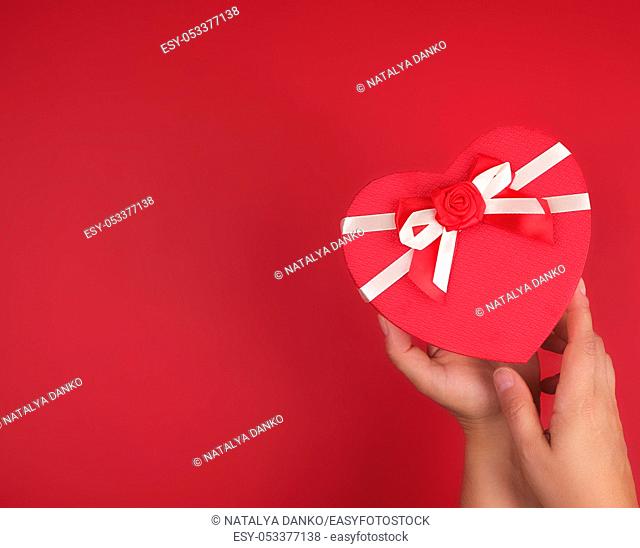 two hands hold a paper closed red box in the shape of a heart on a red background, festive backdrop