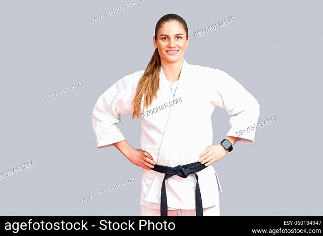 Happy cheerful beautiful blonde young athletic woman in white kimono, black belt standing, holdind hands on waist, toothy smiling and looking at camera